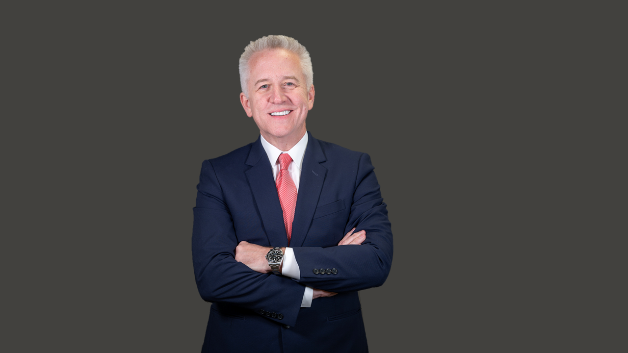 Stephen Toups wears a navy suit and stands in front of a dark grey wall with his arms crossed. 