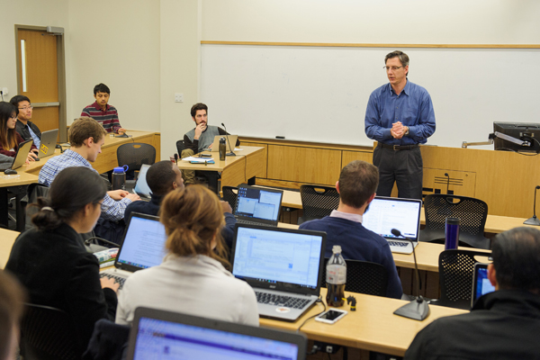 Students in classroom with laptops open, professor is teaching.